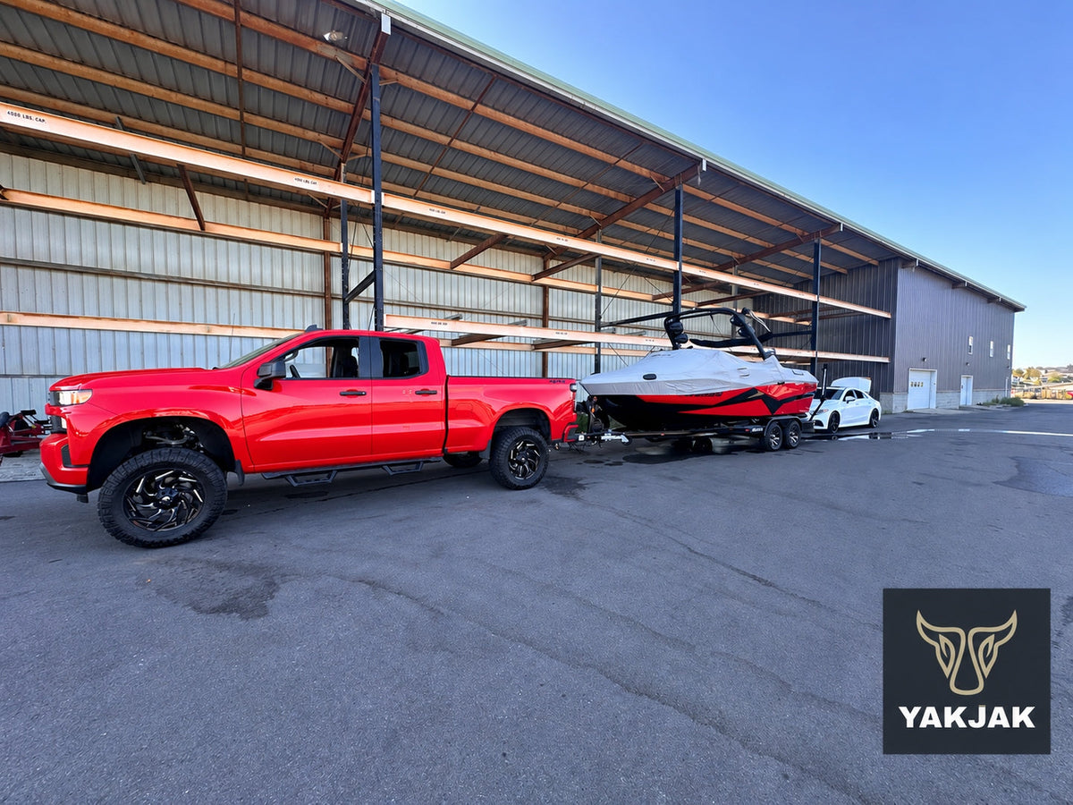 Red pickup towing a covered red-and-black boat on a trailer beside a storage building, with a YakJak logo overlay.